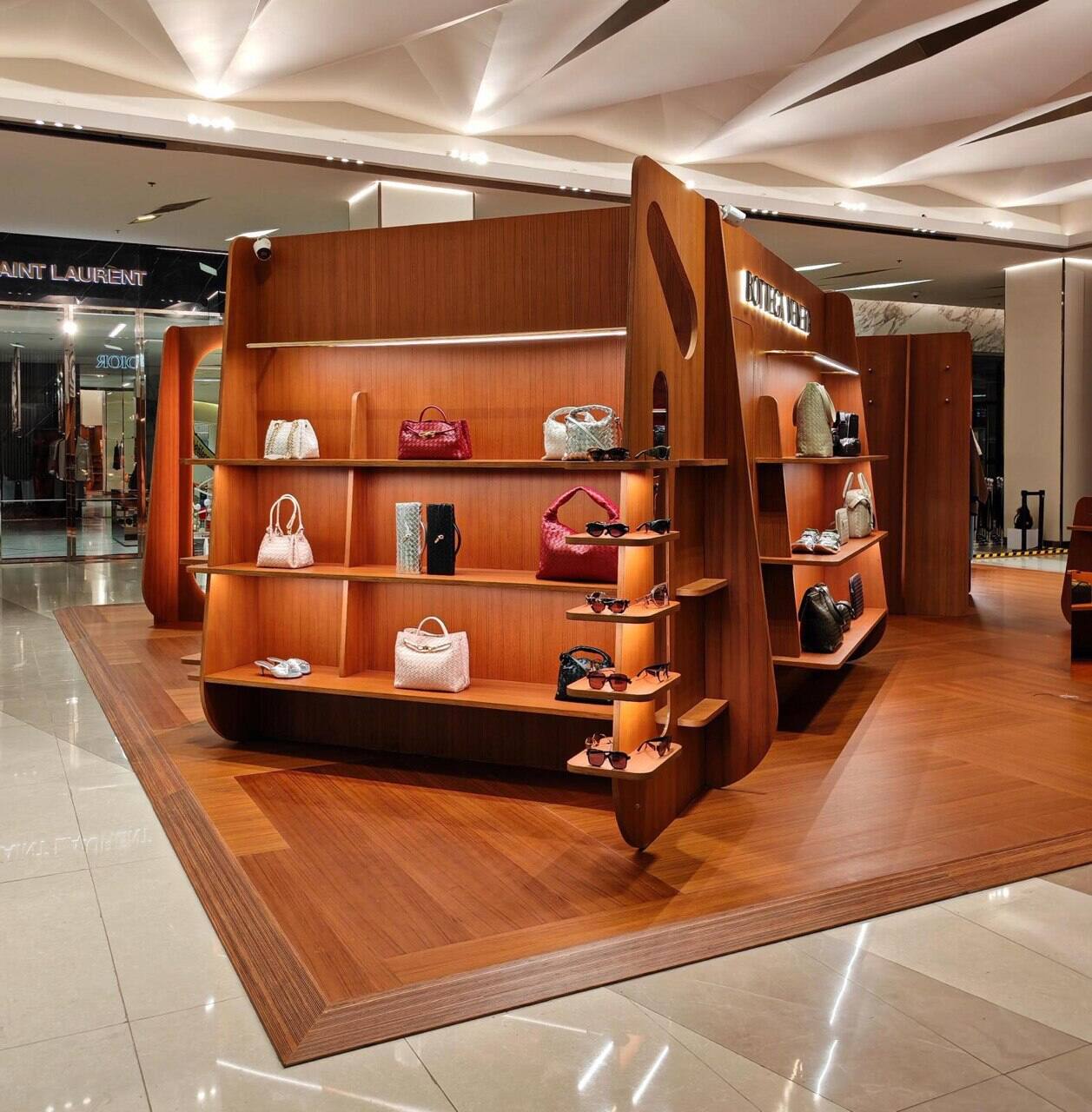 Sculptural timber display island holding handbags and sunglasses beneath a faceted white ceiling