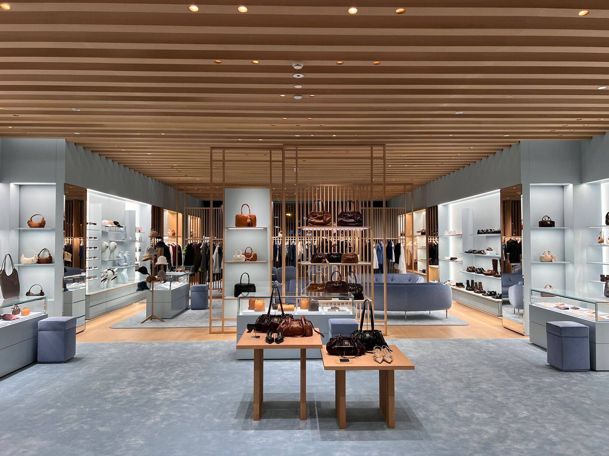 Miu Miu Bangkok store interior with a raked timber batten ceiling, backlit wall vitrines and low timber display tables